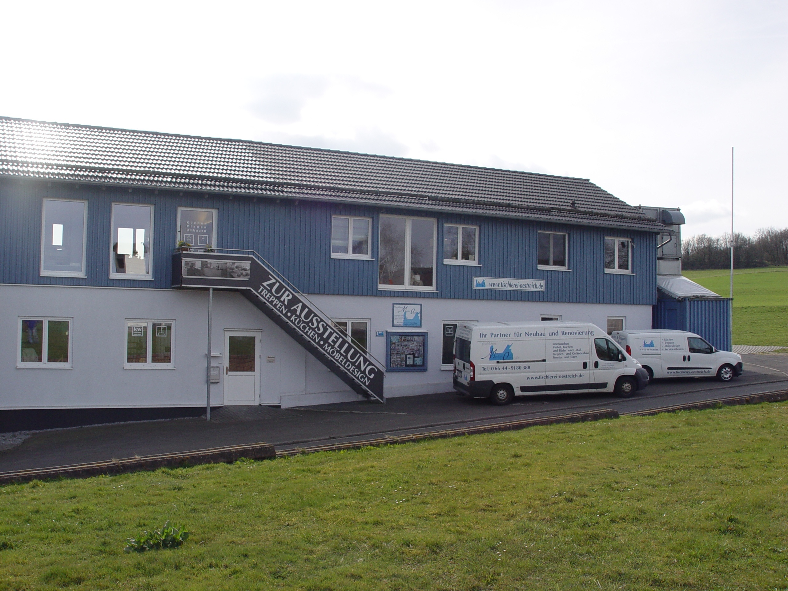 Zweistöckiges Firmengebäude mit Außentreppe und zwei weißen Lieferwagen davor.