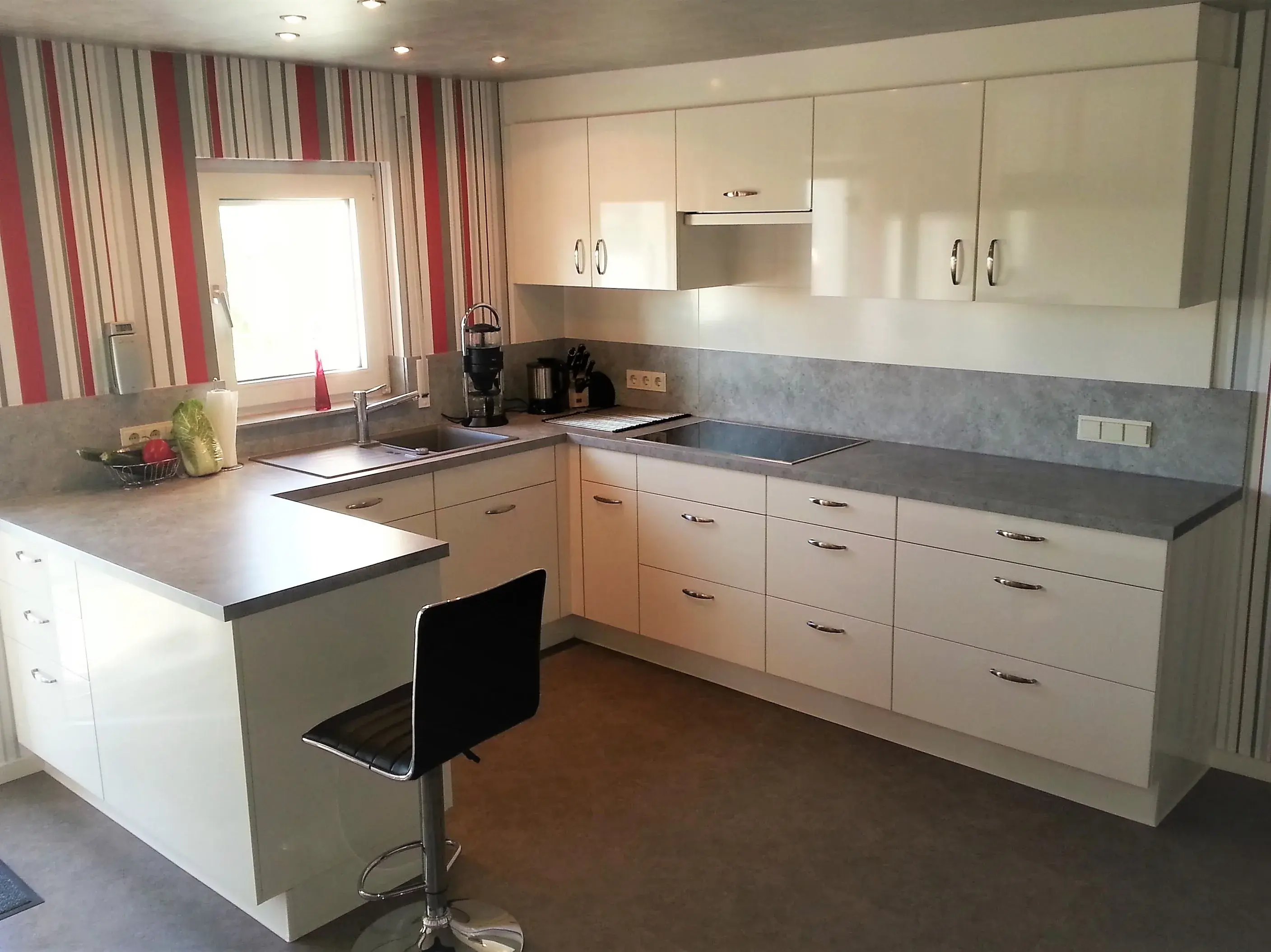 Moderne weiße Einbauküche mit Barhocker und rot-weiß gestreifter Tapete.