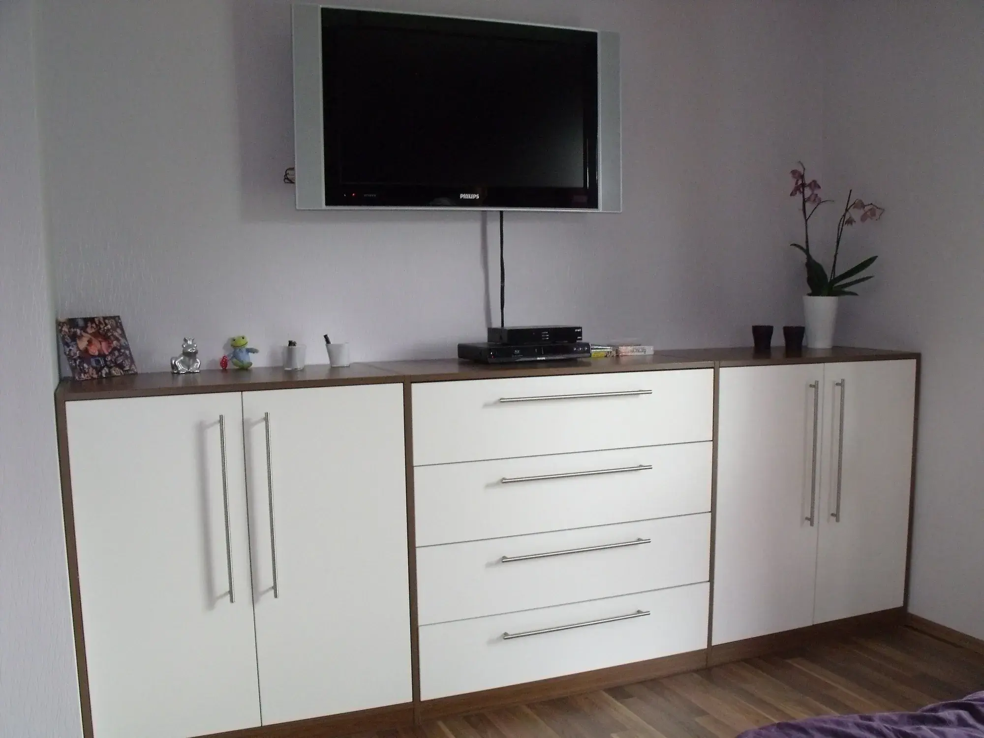 Weiße Kommode mit TV an der Wand, dekoriert mit Pflanzen und Kleinigkeiten.