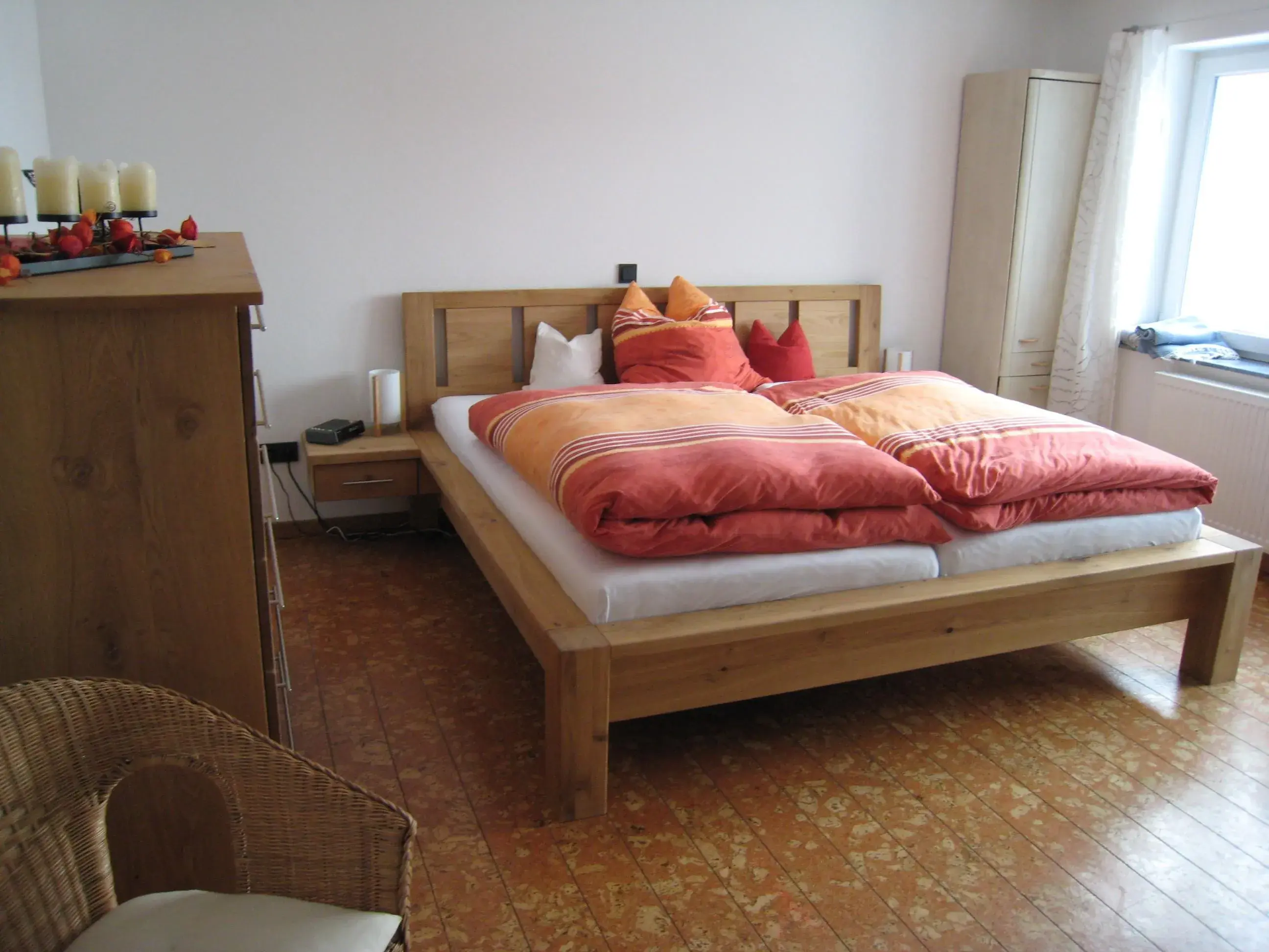 Schlafzimmer mit Holzbett, orangener Bettdecke und Tageslicht durchs Fenster.