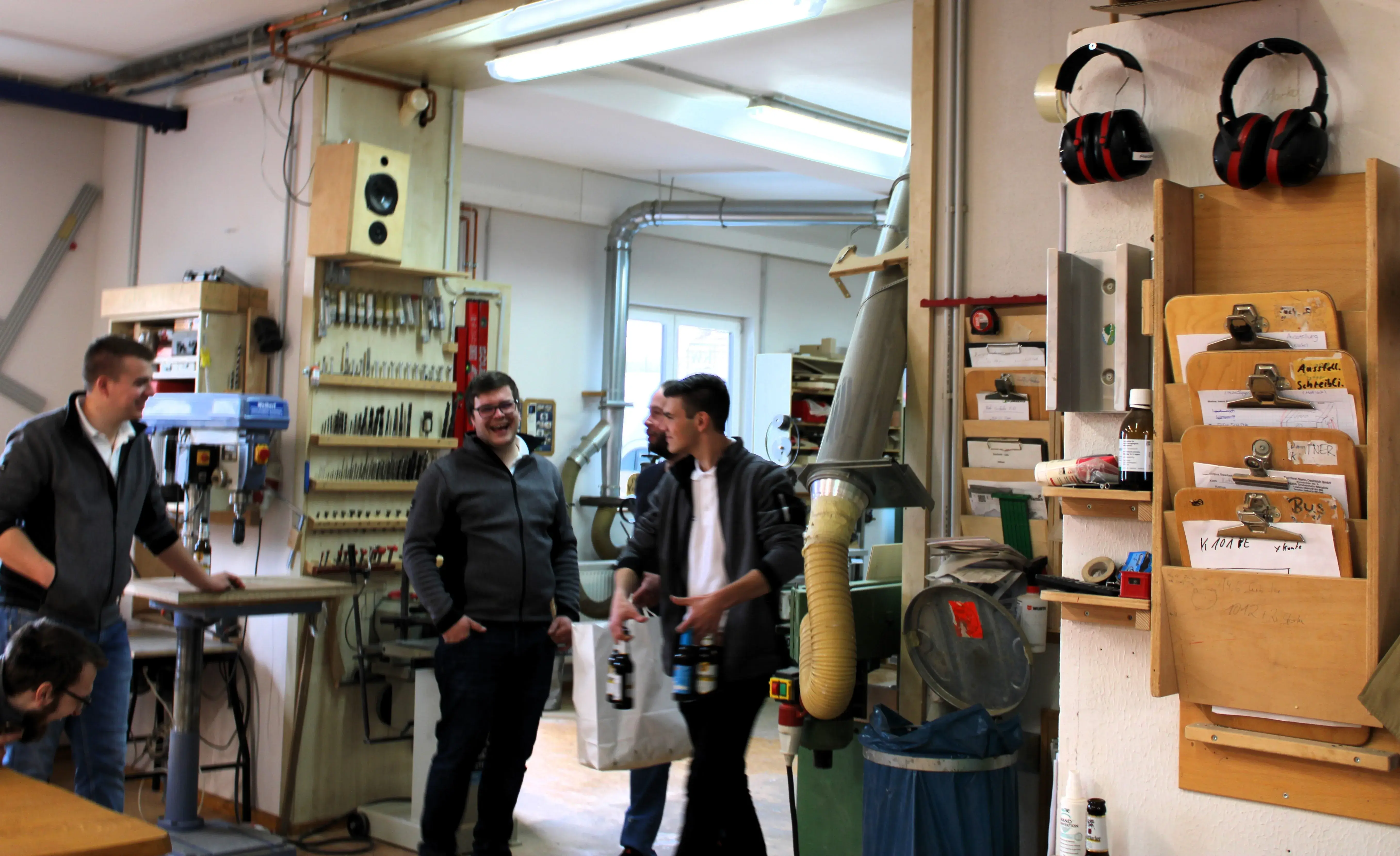 Vier Männer unterhalten sich in einer Werkstatt mit Werkzeugen und Maschinen im Hintergrund.