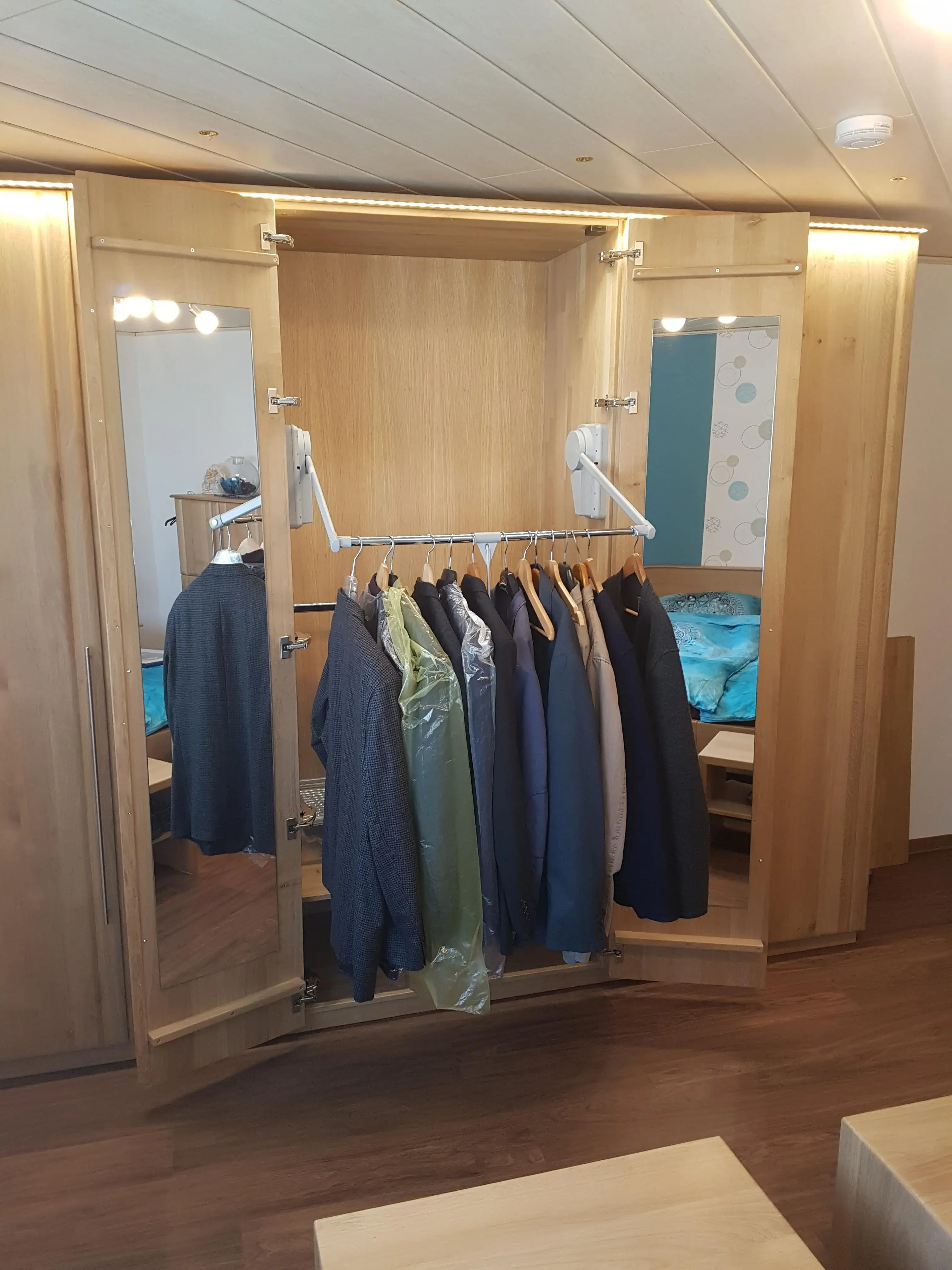 Offener Holzkleiderschrank mit Spiegeln und mehreren hängenden Hemden und Jacken.