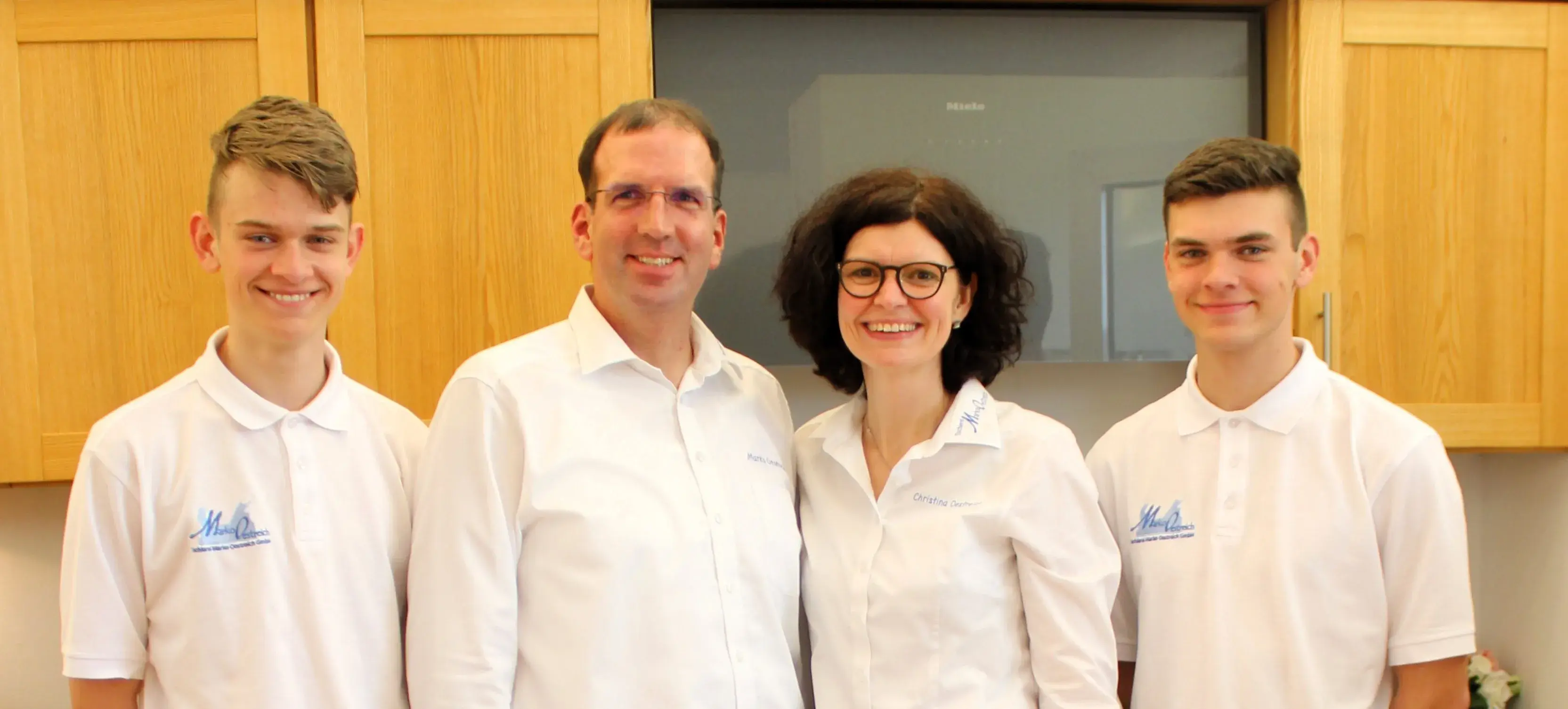 Vier Personen in weißen Hemden lächeln vor Holzschränken in einer Küche.