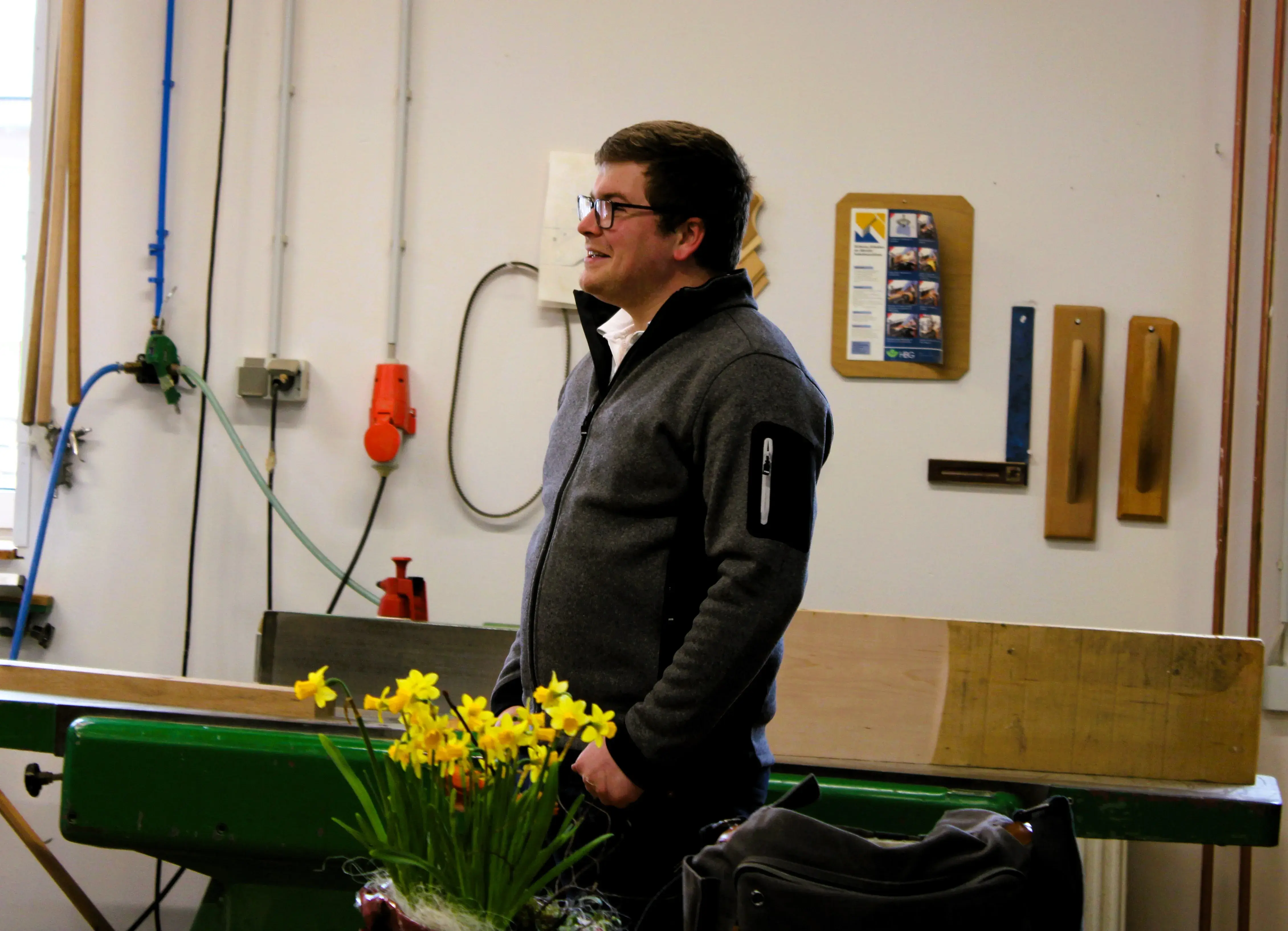 Mann steht in Werkstatt neben gelben Blumen, trägt Brille und dunkle Jacke.