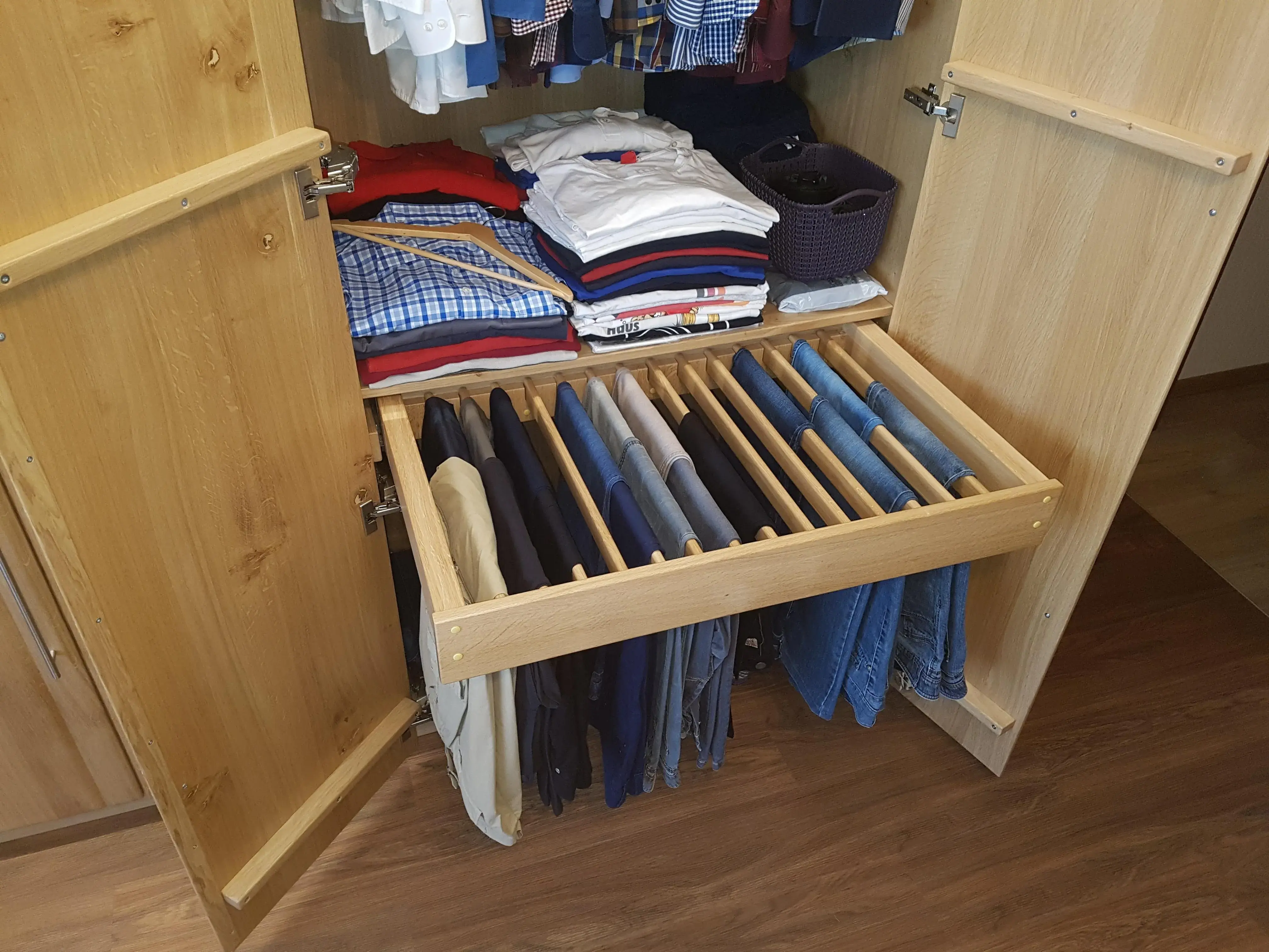 Holzschrank mit Hosen auf Hosenbügeln und gefalteten Hemden auf Regalböden.